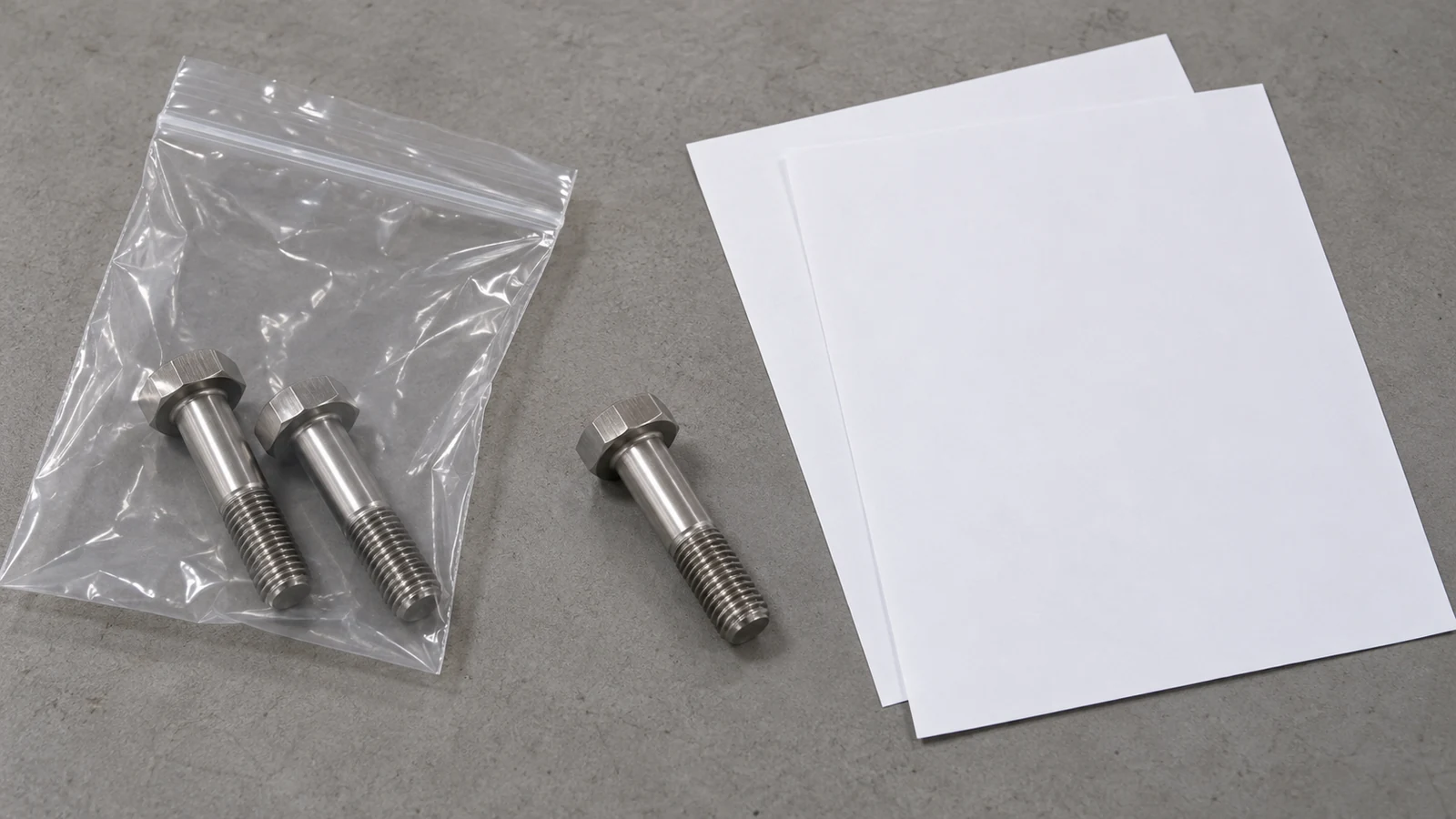 Three visually similar partially threaded hex bolts, clear unlabelled packaging and two blank paper sheets arranged on the same neutral receiving table.